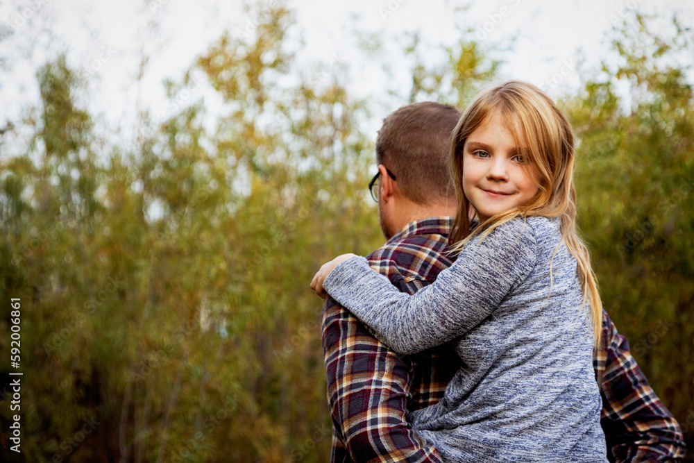 Close-up of a father giving his young daughter a piggyback ride outdoors, the girl looking back over her shoulder at the camera; Edmonton, Alberta, Canada Close-up of a father giving his young daughter a piggyback ride outdoors, the girl looking back over her shoulder at the camera; Edmonton, Alberta, Canada