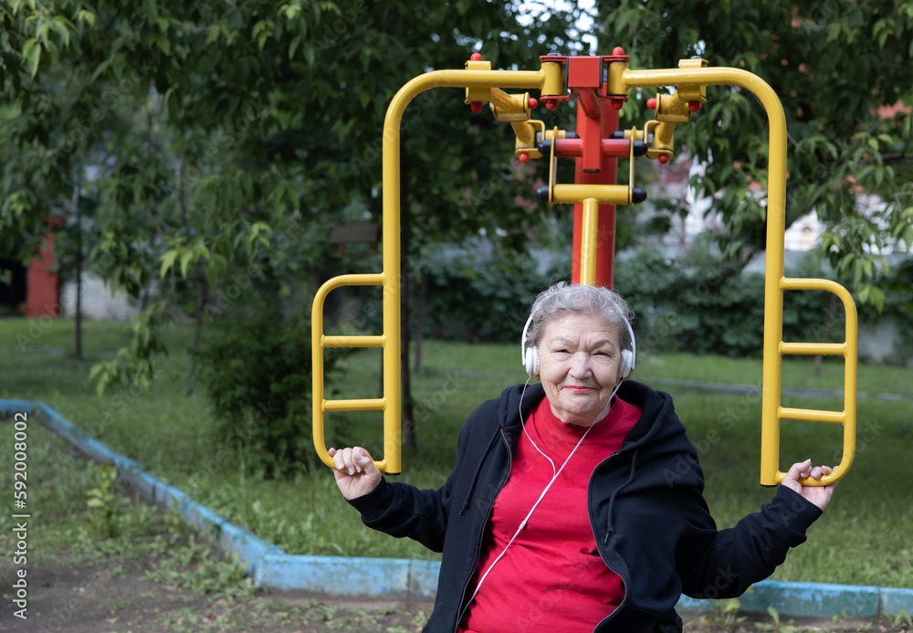 Obraz premium Smiling elderly woman in headphones is doing exercises on simulator, trainer outdoors in the yard. Active life of pensioners. Adaptation of pensioners in the modern world. Prevention of mental illness
