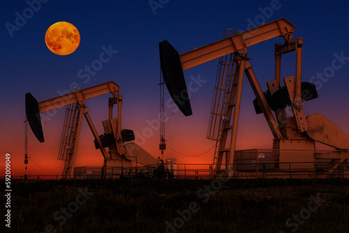 Glowing colourful pumpjacks at sunrise with glowing sky and full moon in the background, West of Airdrie; Alberta, Canada