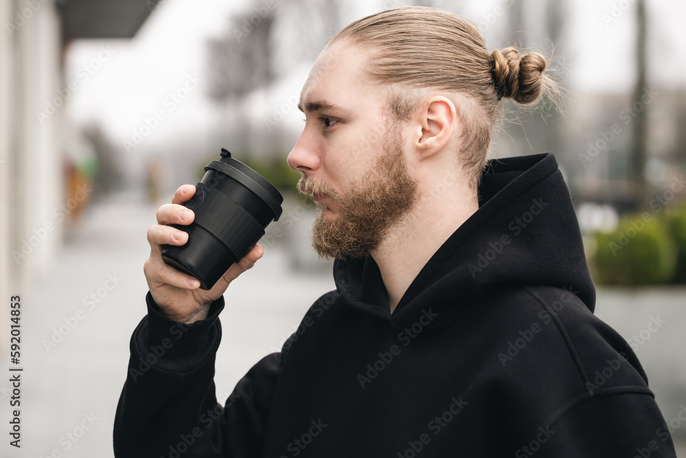 A young bearded man drinks from a stylish black bamboo reusable cup outdoors. Reusable eco friendly sustainable handy bamboo cup zero waste, no plastic concept. A young bearded man drinks from a stylish black bamboo reusable cup outdoors. Reusable eco friendly sustainable handy bamboo cup zero waste, no plastic concept.