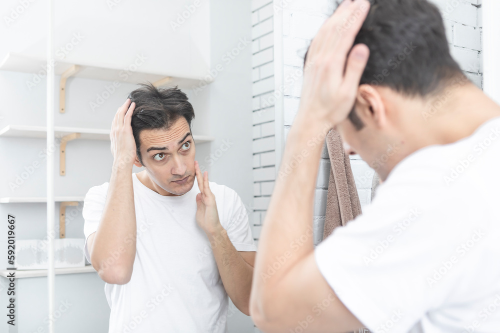 Handsome man looking at mirror and applying moisturizing cream on cheeks in bathroom. Groomed young guy doing skincare morning routine after taking a shower Handsome man looking at mirror and applying moisturizing cream on cheeks in bathroom. Groomed young guy doing skincare morning routine after taking a shower