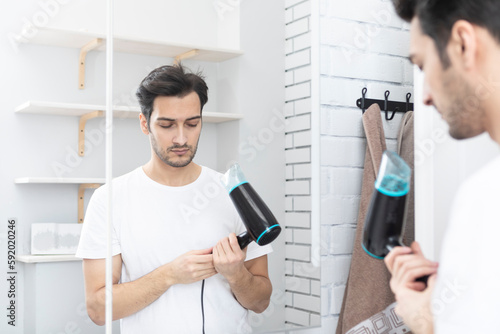 Young handsome man standing in front of the mirror in the bathroom and holding hair dryer. Morning routine and making hair style concept