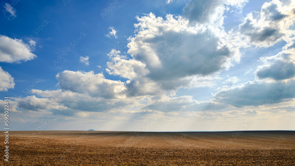 Clouds in the sunny blue sky over the plowed wavy brown-yellowish agricultural field with a little hill in the background. Landscape panorama. 
