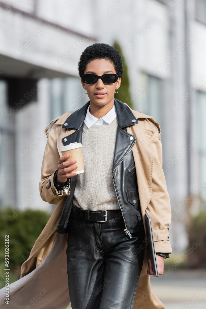 african american businesswoman in trench coat and black leather pants walking with coffee to go and laptop on blurred city street.