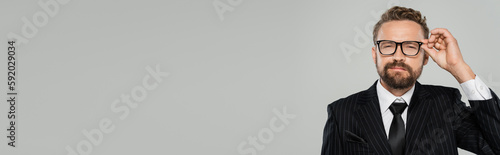 bearded businessman in formal wear adjusting glasses and looking at camera isolated on grey, banner.