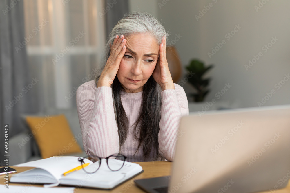 Despaired sad caucasian old lady sitting at table, looking at computer, suffering from headache, menopause, stress Despaired sad caucasian old lady sitting at table, looking at computer, suffering from headache, menopause, stress