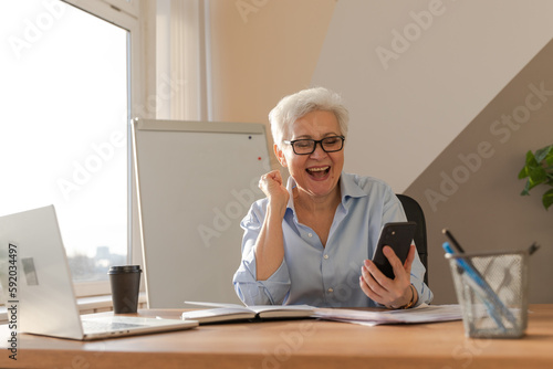 Confident middle aged senior woman euphoric winner with smartphone. Older mature lady looking at cell phone reading great news getting good result winning online bid feeling amazed. Winning gesture