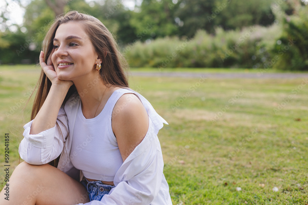 Nice looking woman is sitting on the grass in park, hold her head and dreaming. She is relaxing, enjoying of the day. Girl look sensual and tenderness. Long hair female wear white shirt and top. Nice looking woman is sitting on the grass in park, hold her head and dreaming. She is relaxing, enjoying of the day. Girl look sensual and tenderness. Long hair female wear white shirt and top.