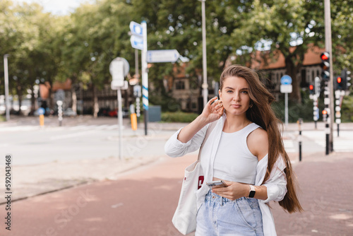 Beautiful young caucasian woman looking at camera walking outdoors. Brunette wears white shirt, blue denim jeans short and shopping bag. Rest time. Long brunette hair woman look at camera, touch face.