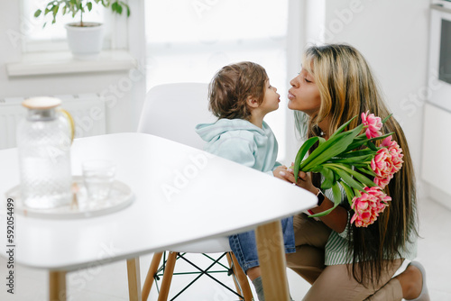 A small child congratulates his mom in the kitchen and gifts a flowers