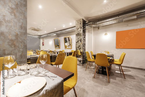 Dining room of a restaurant with tables, with cutlery, crockery, glassware and assembled chairs upholstered in yellow fabric and exposed air conditioning pipes
