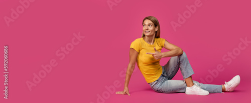 Positive middle aged european blonde lady sitting on floor pointing finger at empty space
