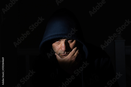 A man wearing a hood is in a dark room, holding his chin and displaying a secretive expression while scheming something sinister.