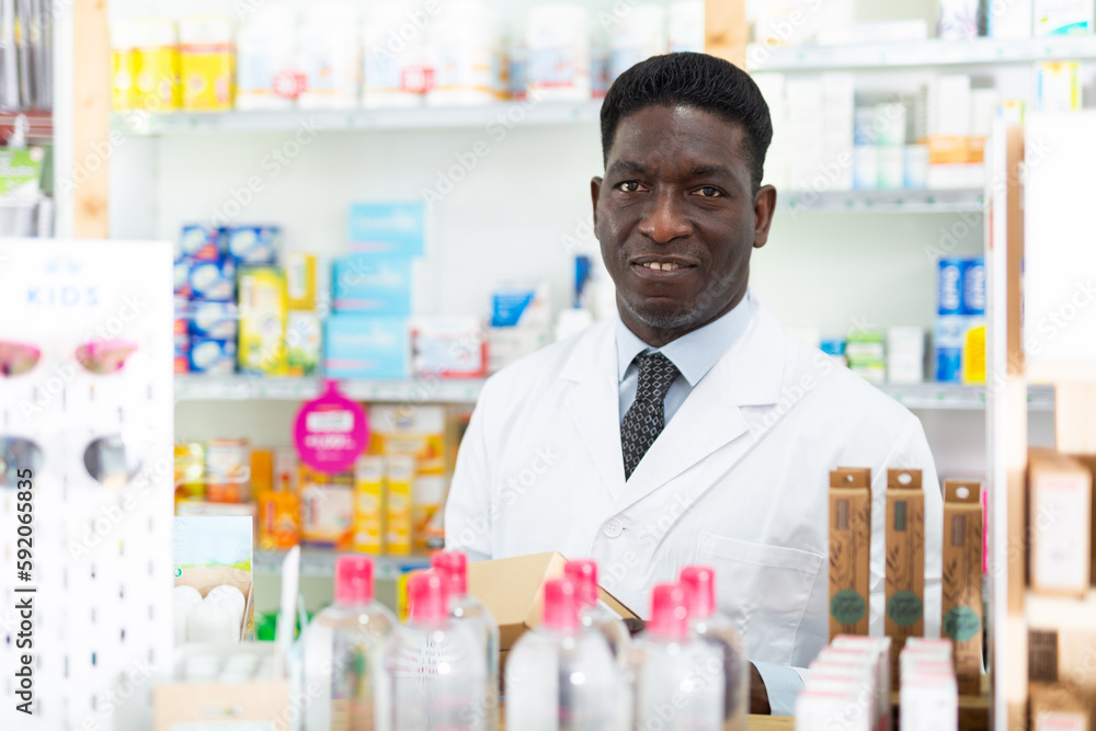 African-american male pharmacist in gown standing behind counter in drugstore