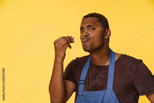 Stylish African American cook wearing blue apron sends kiss with his fingers in Italian manner, against bright yellow background with copy space for text or product placement.