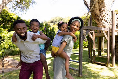 Happy african american parents piggybacking son and daughter while standing in park, copy space