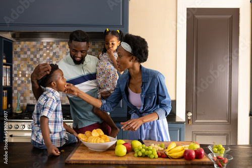 African american mother feeding fresh fruits to children and husband while standing in kitchen