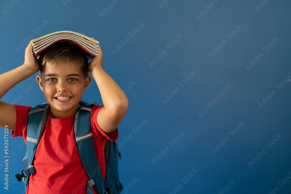 Portrait of smiling caucasian boy carrying books on head while standing over blue background Portrait of smiling caucasian boy carrying books on head while standing over blue background