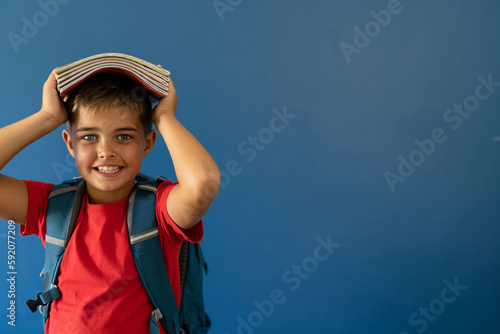 Portrait of smiling caucasian boy carrying books on head while standing over blue background