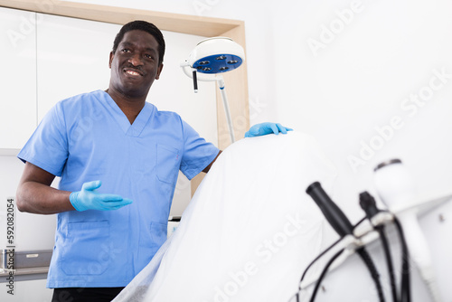 Smiling confident man beautician wearing blue overall posing in clinic of esthetic medicine