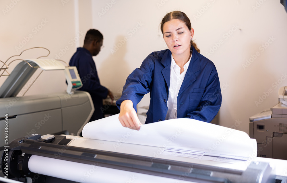 Concentrated young female in a blue robe uniform using plotter while printing a banner in the typography
