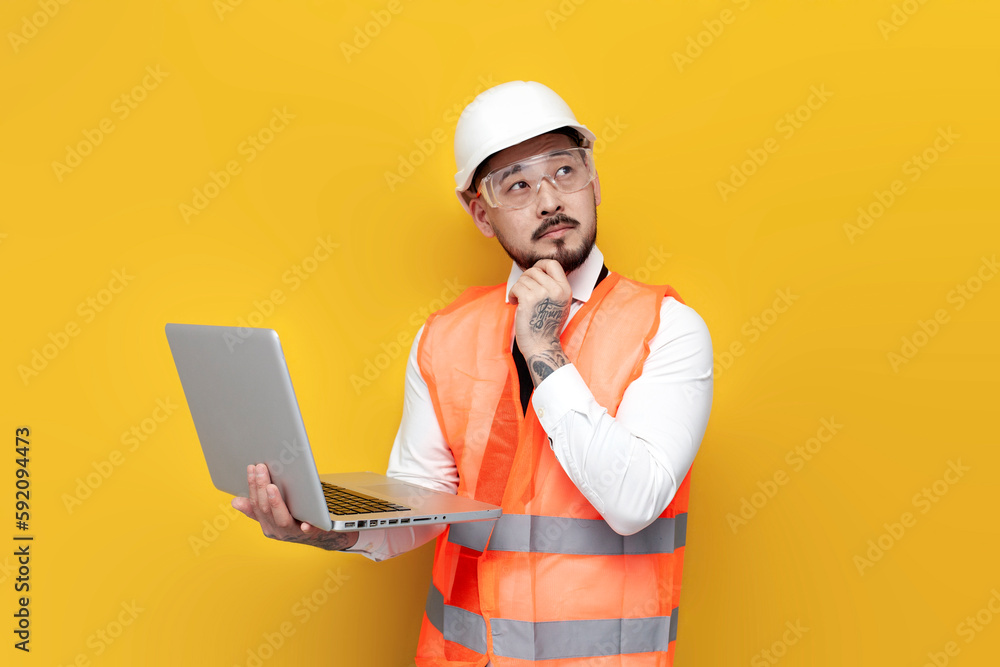 asian pensive foreman in uniform holds laptop on yellow isolated background and thinks, korean civil engineer asian pensive foreman in uniform holds laptop on yellow isolated background and thinks, korean civil engineer