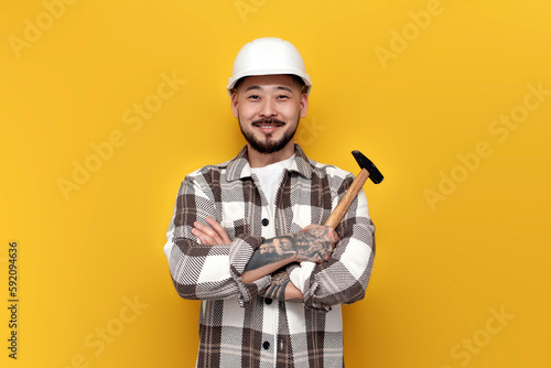 asian builder in hard hat and shirt holds hammer on yellow isolated background, korean worker constructor