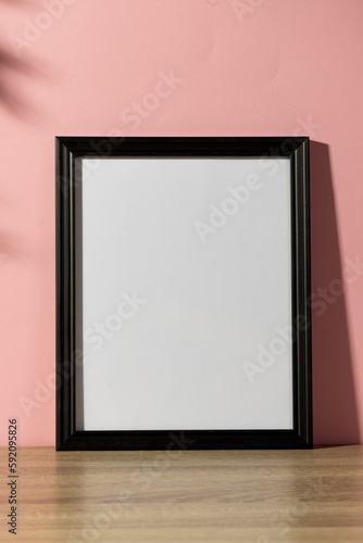 Vertical of black empty frame with copy space on table against pink wall and shadow of leaves