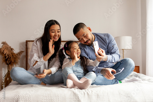 asian family sit on the bed and communicate via video call, little korean girl with mom and dad takes selfie