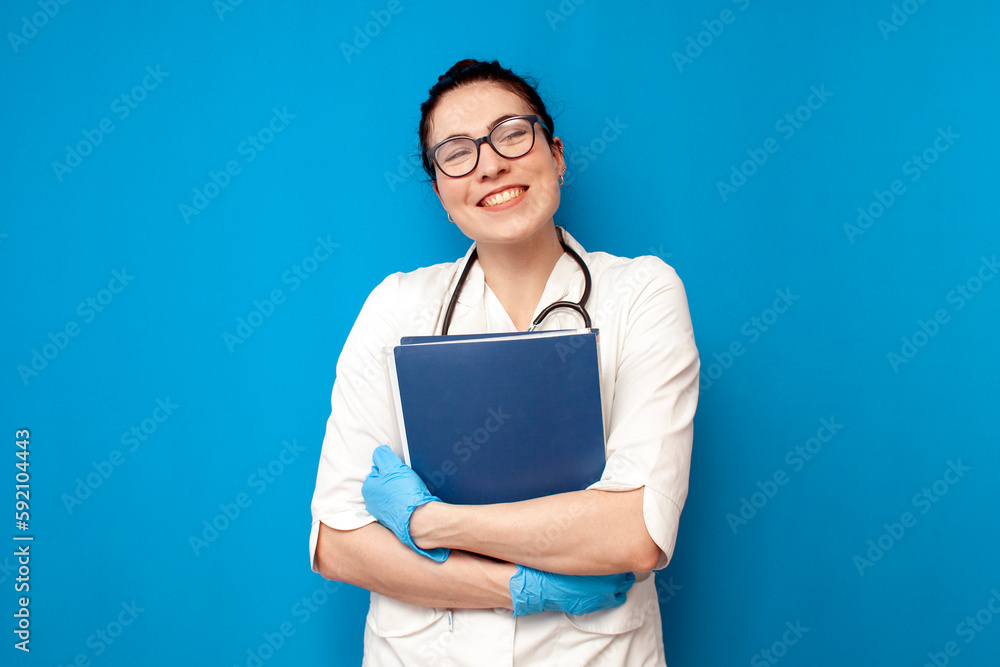 female doctor student in a medical gown holds documents and papers on a blue isolated background, a girl intern nurse female doctor student in a medical gown holds documents and papers on a blue isolated background, a girl intern nurse