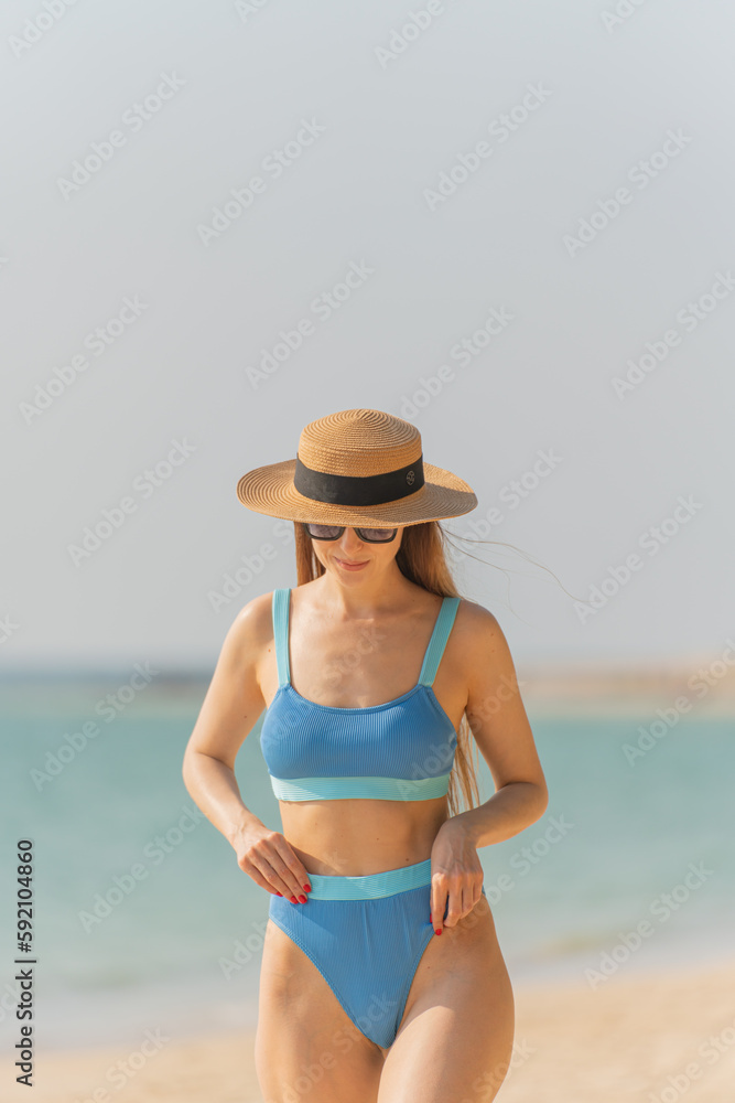 Beautiful caucasian blonde woman in bikini with beach hat and sunglasses wandering by the beach Beautiful caucasian blonde woman in bikini with beach hat and sunglasses wandering by the beach