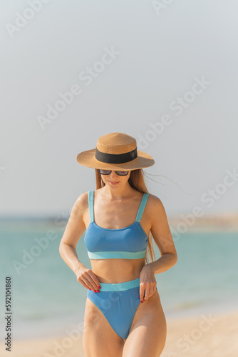Beautiful caucasian blonde woman in bikini with beach hat and sunglasses wandering by the beach