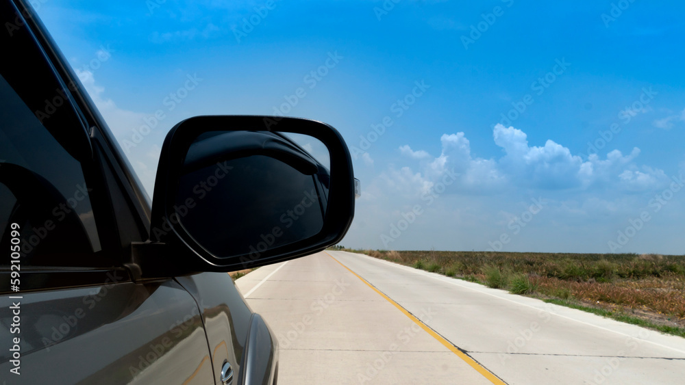 Beside of gray car driving on concrete road. Can view mirror wing of car. Beside with slope of concrete road and blurred of open space under blue sky.
