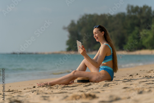 Beautiful caucasian woman using digital tablet at the beach. Female digital nomad working from the beach using her digital tablet. Sexy white woman in bikini. Workcation. © asean studio