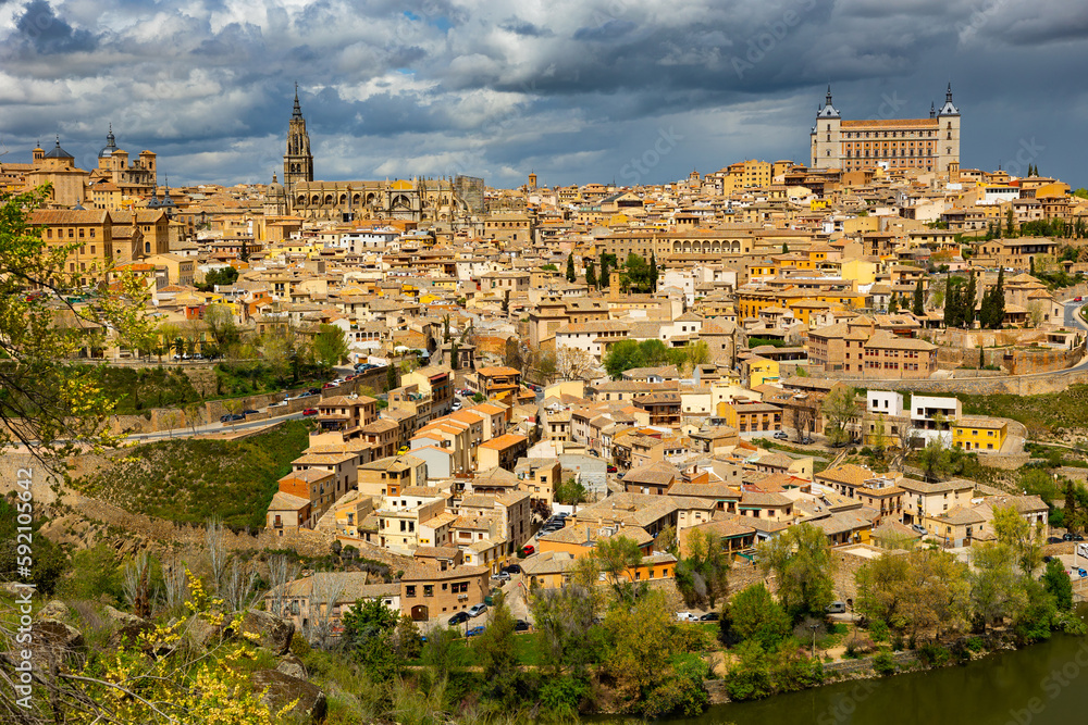 View of historic area of Toledo city on banks of Tagus River overlooking belfry of Primatial Cathedral of Saint Mary and Alcazar fortress in cloudy spring day, Spain