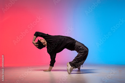young guy dancer in black clothes break dancing in neon lighting, man performs hip hop movement in red blue club