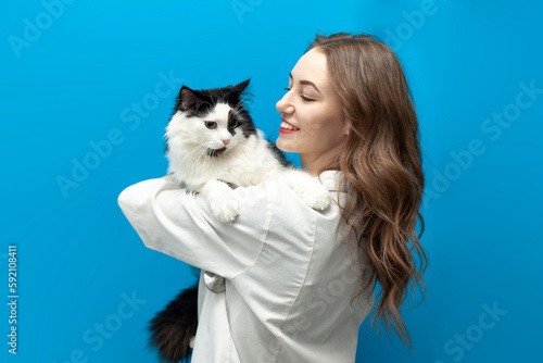 young girl veterinarian in uniform holds black and white cat on blue isolated background, doctor in medical coat
