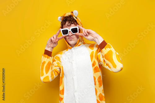 Photography young joyful guy in funny baby giraffe pajamas and glasses is dancing on yellow