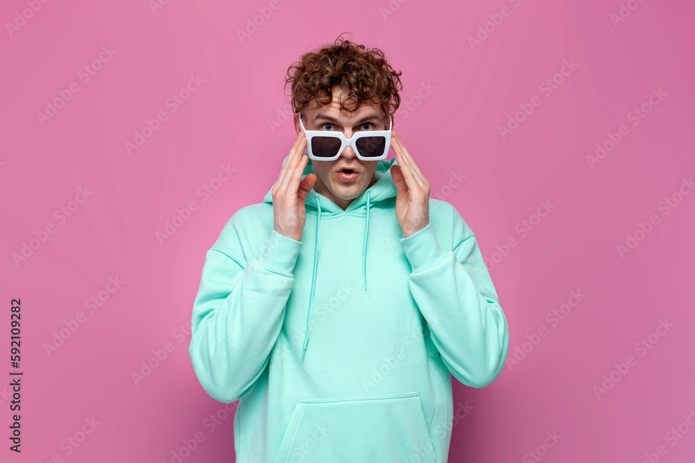 young curly shocked guy in mint hoodie with sunglasses looks in surprise on pink isolated background young curly shocked guy in mint hoodie with sunglasses looks in surprise on pink isolated background