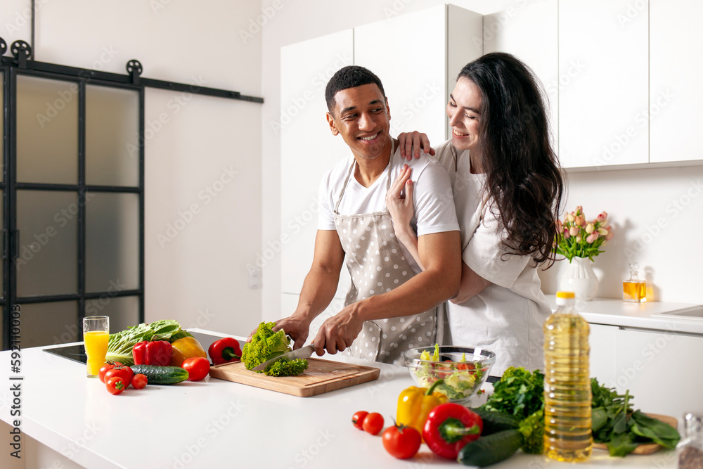 young happy multiracial couple preparing veggie vegetable and greens salad in white modern kitchen