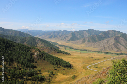 A narrow asphalt road windingly crosses a valley sandwiched in the mountains on a sunny summer day.