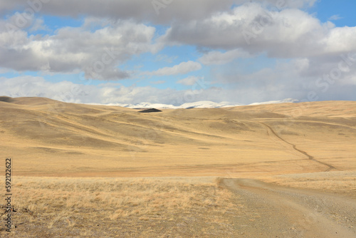 A narrow dirt road descends from the top of the hill into the autumn arid steppe to meet high snow-capped mountains on a clear autumn day.