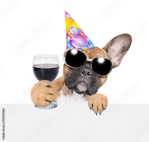 French bulldog puppy wearing sunglasses and party cap looks above empty white banner and holds glass of red wine. isolated on white background © Ermolaev Alexandr
