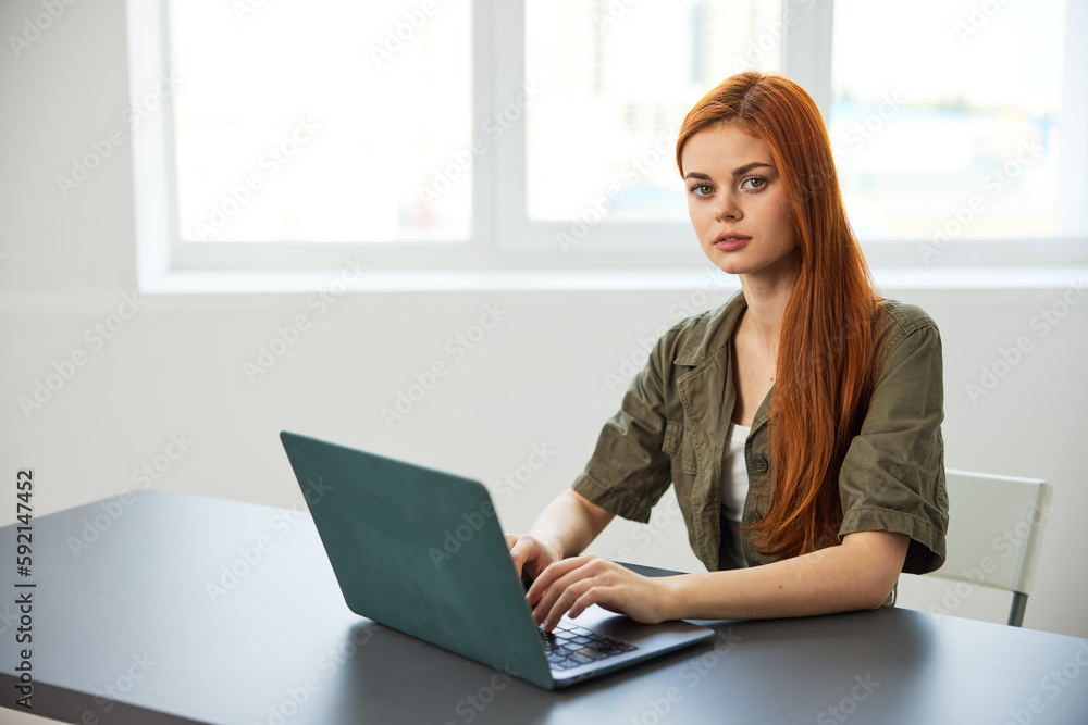 red-haired woman typing in a laptop sitting at a table in the office near the window working full time red-haired woman typing in a laptop sitting at a table in the office near the window working full time