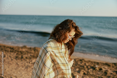 a woman in large glasses wraps herself in a blanket walking along the sea coast