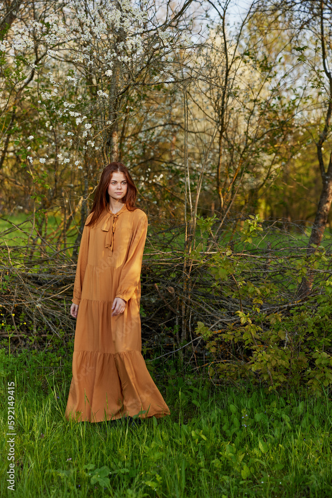 an elegant, sophisticated woman poses relaxed standing near a wicker fence at the dacha in a long orange dress an elegant, sophisticated woman poses relaxed standing near a wicker fence at the dacha in a long orange dress