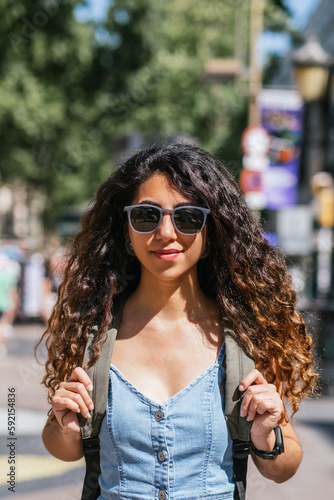 portrait of a beautiful dark haired young woman wearing denim dress. She wears sunglasses and a backpack and looks to camera with confidence