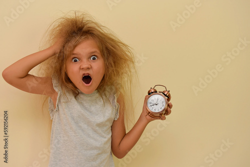 A sleepy child yawns with an alarm clock in his hands. Early rise to school or kindergarten. The kid was awakened early in the morning with tousled hair. The little girl did not get enough sleep.
