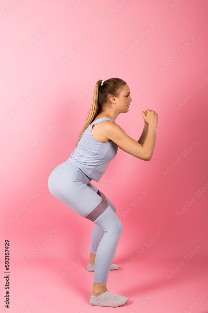 Young healthy Girl in gray sportswear is doing stretches and warm-up exercises with a rubber band resistance. Isolated on pink background. Young healthy Girl in gray sportswear is doing stretches and warm-up exercises with a rubber band resistance. Isolated on pink background.