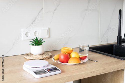 A plate of fresh fruit on the kitchen table, a kitchen scale and a glass of water near the sink. The concept of healthy and proper nutrition